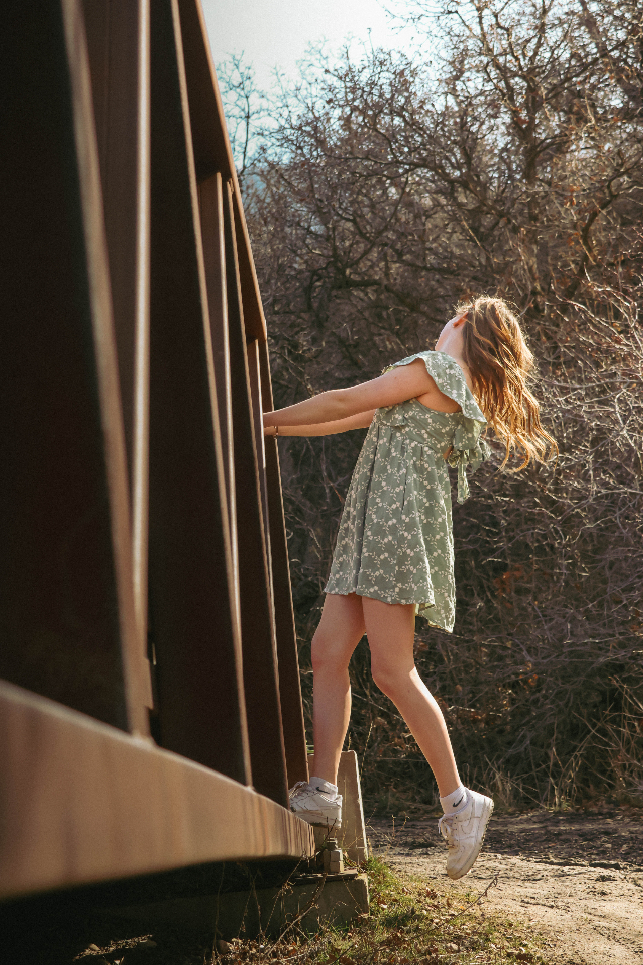 Girl in green floral dress leaning playfully off a wooden bridge in golden light